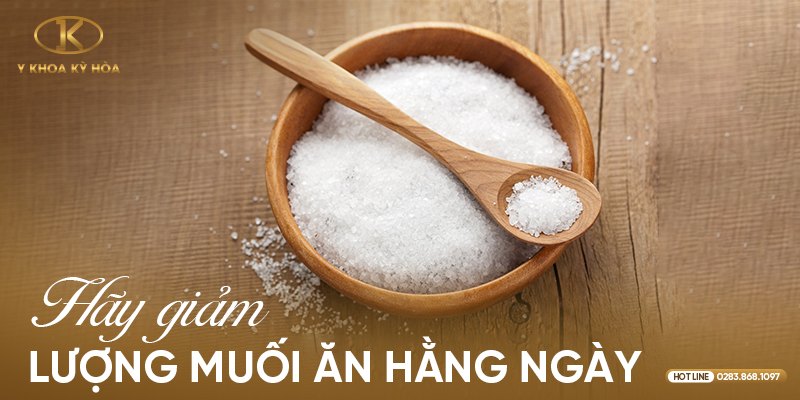 Chén muối trắng và muỗng gỗ trên mặt bàn gỗ kèm dòng chữ Hãy giảm lượng muối ăn hằng ngày